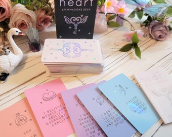 Könnte beinhalten: Ein Deck Affirmationskarten mit einer schwarzen Box, auf der der Text "ALIGN: heart AFFIRMATIONS DECK" steht. Die Karten haben ein holografisches Design mit einem Schlüssel und Flügeln. Die Karten liegen auf einer weißen Holzoberfläche mit rosa Blumen und einer weißen Schwanenfigur.