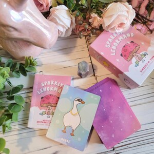 May include: A pink and white deck of cards called "Sparkly Lenormand" with a mushroom illustration on the box. The cards feature a pink and purple galaxy design with a goose card showing a goose wearing sunglasses.