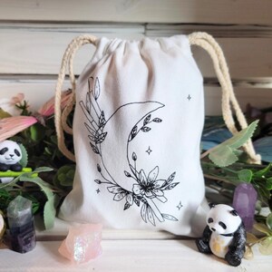 May include: A white cotton drawstring bag with a black and white illustration of a crescent moon, flowers, and stars. The bag is perfect for storing small items like crystals, jewelry, or other trinkets.