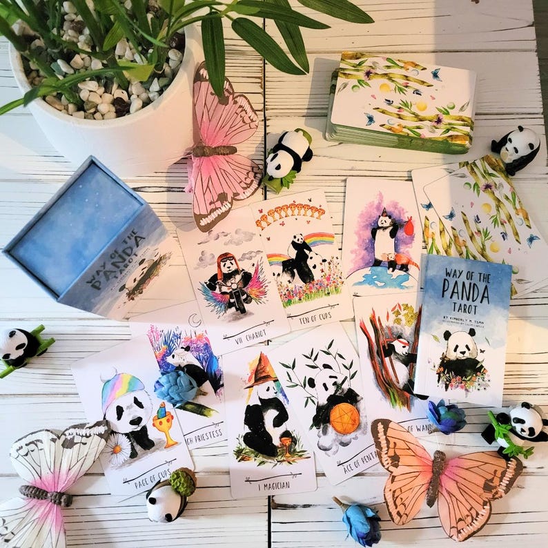 May include: A set of tarot cards featuring illustrations of pandas in various poses and settings. The cards are laid out on a wooden table with a box labeled "Way of the Panda Tarot".