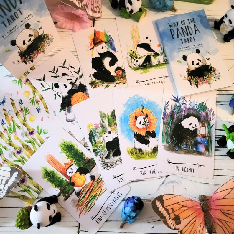 May include: A set of Way of the Panda Tarot cards featuring watercolor illustrations of pandas in various poses and settings. The cards are laid out on a white wooden surface with a few small decorative items.