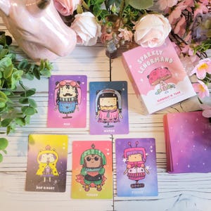 May include: A set of colorful cards with illustrations of people in different outfits. The cards are part of a deck called "Sparkly Lenormand" by Kimberly M. Tran. The cards are laid out on a white wooden surface with pink flowers and a pink unicorn figurine.