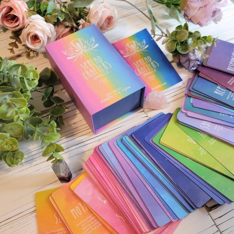 K&ouml;nnte beinhalten: Ein Deck Spread Crafters Oracle Karten mit einem Regenbogen-Farbverlauf als Hintergrund. Die Karten sind aufgef&auml;chert, um die verschiedenen Farben und Designs zu zeigen. Die Schachtel f&uuml;r die Karten ist lila mit einem Regenbogen-Farbverlauf und dem Text "Spread Crafters Oracle" in Wei&szlig;.