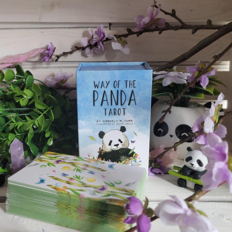 May include: A deck of Way of the Panda Tarot cards with a blue box featuring a black and white panda illustration. The box is sitting on a white surface with a small panda figurine and a potted plant with pink flowers.