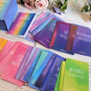 K&ouml;nnte beinhalten: Ein Deck Spread Crafters Oracle-Karten mit einem Handbuch. Die Karten haben einen regenbogenfarbenen Verlaufshintergrund mit einem galaktischen Muster. Die Karten zeigen W&ouml;rter wie "Verantwortungsbewusst", "Integrieren", "Schattenarbeit" und "Fokusbereiche".