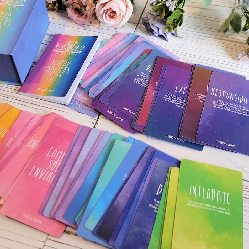 K&ouml;nnte beinhalten: Ein Deck Spread Crafters Oracle-Karten mit einem Handbuch. Die Karten haben einen regenbogenfarbenen Verlaufshintergrund mit einem galaktischen Muster. Die Karten zeigen W&ouml;rter wie "Verantwortungsbewusst", "Integrieren", "Schattenarbeit" und "Fokusbereiche".