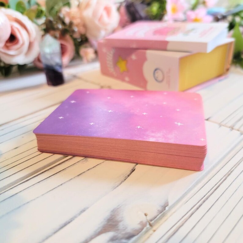 May include: A deck of pink and purple affirmation cards with a galaxy design and white stars. The cards are stacked on a white wooden surface.