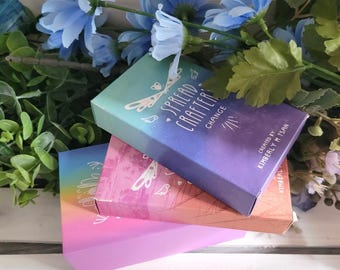 Könnte beinhalten: Drei farbenfrohe Schachteln mit "Spread Crafter Change" Orakelkarten mit einem Libellen-Design. Die Schachteln sind übereinander gestapelt, wobei die oberste Schachtel einen hellblauen und violetten Farbverlauf hat, die mittlere Schachtel einen rosa und orangenen Farbverlauf und die unterste Schachtel einen violetten und rosa Farbverlauf.