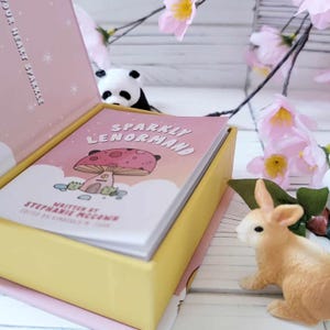 May include: A pink and yellow box with a deck of cards inside. The cards have a pink and white background with a mushroom illustration and the text "Sparkly Lenormand". The box is open and the cards are visible. The text "Written by Stephanie McCann" is also visible on the cards.