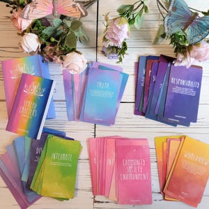 K&ouml;nnte beinhalten: Ein Set farbenfroher Orakelkarten mit Aquarellhintergr&uuml;nden und wei&szlig;em Text. Die Karten liegen auf einer wei&szlig;en Holzoberfl&auml;che mit einem Begleitbuch und Blumen. Die Karten enthalten W&ouml;rter wie "Wahrheit", "Integrieren", "Gemeinschaft, Gesellschaft, Umwelt" und "Weltbild".