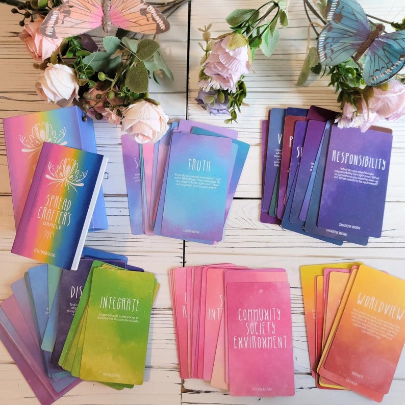 K&ouml;nnte beinhalten: Ein Set farbenfroher Orakelkarten mit Aquarellhintergr&uuml;nden und wei&szlig;em Text. Die Karten liegen auf einer wei&szlig;en Holzoberfl&auml;che mit einem Begleitbuch und Blumen. Die Karten enthalten W&ouml;rter wie "Wahrheit", "Integrieren", "Gemeinschaft, Gesellschaft, Umwelt" und "Weltbild".