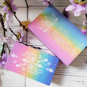 K&ouml;nnte beinhalten: Ein Deck Orakelkarten namens "Spread Crafters Oracle" mit einem Regenbogen-Farbverlauf im Hintergrund und einem wei&szlig;en Schmetterlingsdesign. Die Schachtel ist rosa mit einem Regenbogen-Farbverlauf und dem Text "Spread Crafters Oracle" in Wei&szlig;. Der Text "Erstellt von Kimberly M. Tsan" ist in Wei&szlig; auf der Schachtel.