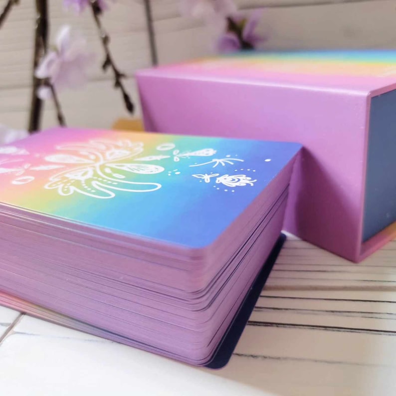 K&ouml;nnte beinhalten: Ein Stapel Karten mit einem Regenbogen-Farbverlaufshintergrund und wei&szlig;en floralen Designs. Die Karten befinden sich in einer violetten Schachtel mit einem Regenbogen-Farbverlaufsdeckel.