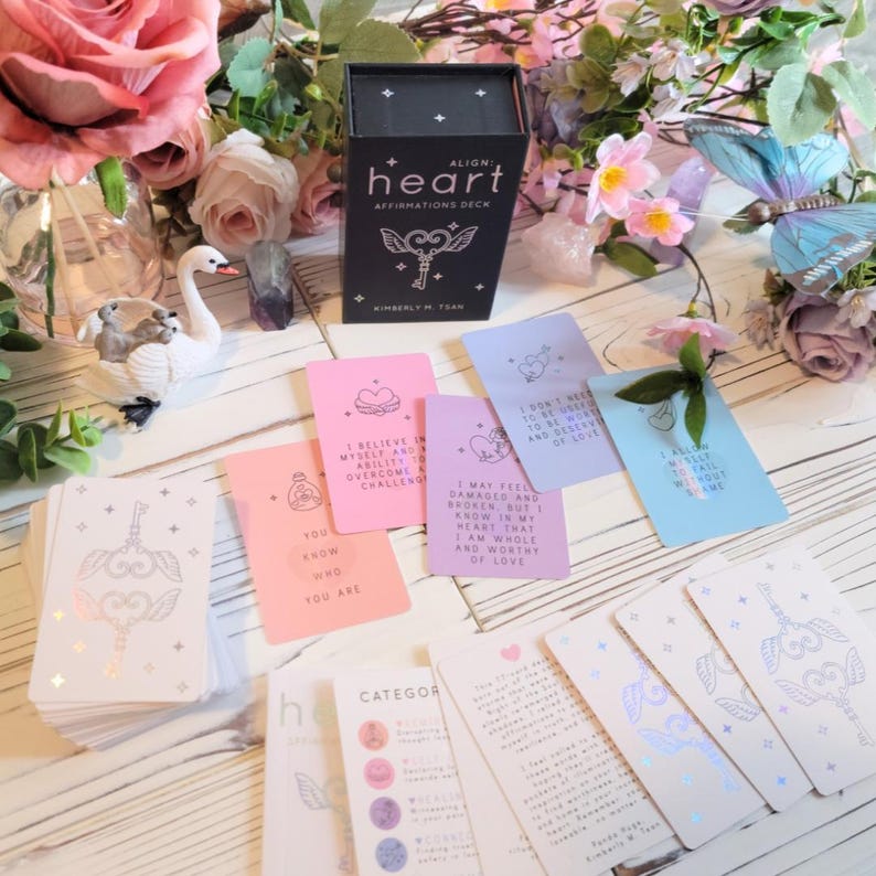 May include: A deck of affirmation cards with a black box that says "Align Heart Affirmations Deck" and a keyhole design. The cards are pink, purple, blue, and white with gold foil accents. The cards have positive affirmations written on them.