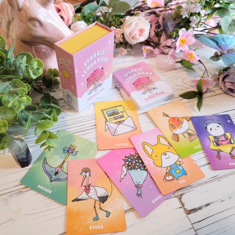 May include: A set of colorful illustrated oracle cards with a pink and yellow box. The box says "Sparkly Lenormand" and the cards feature illustrations of a letter, a fish, a dog, a stork, a bouquet of flowers, an anchor, and a bird.