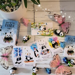 May include: A set of tarot cards featuring illustrations of pandas in various poses and settings. The cards are laid out on a white wooden surface with a book titled "Way of the Panda Tarot" and two butterflies.