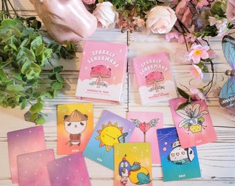 May include: A set of pink and blue illustrated oracle cards with a sparkly pink background. The cards feature cute cartoon animals and symbols. The cards are titled "Sparkly Lenormand" and are designed by Kimberly M. Tear.