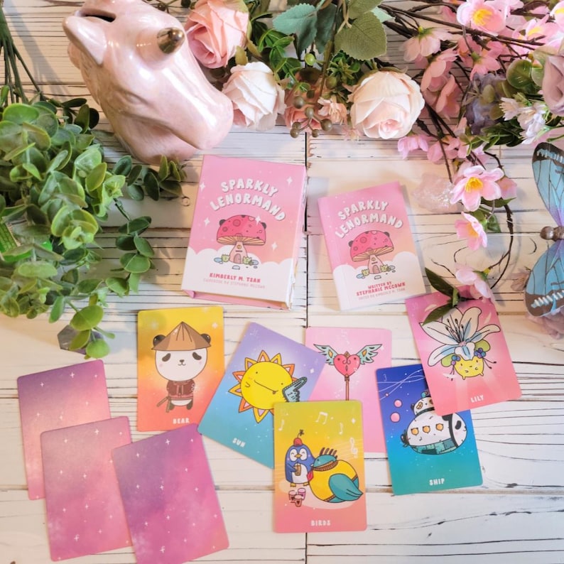 May include: A set of pink and blue illustrated oracle cards with a sparkly pink background. The cards feature cute cartoon animals and symbols. The cards are titled "Sparkly Lenormand" and are designed by Kimberly M. Tear.