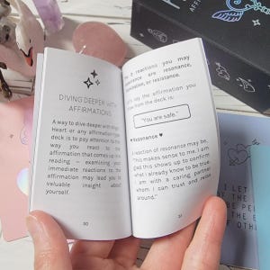 May include: A small white book with a pink cover and a black cover. The book is open to a page with the title "Diving Deeper with Affirmations". The text on the page describes how to use affirmations to connect with your heart. The page also includes the affirmation "You are safe."