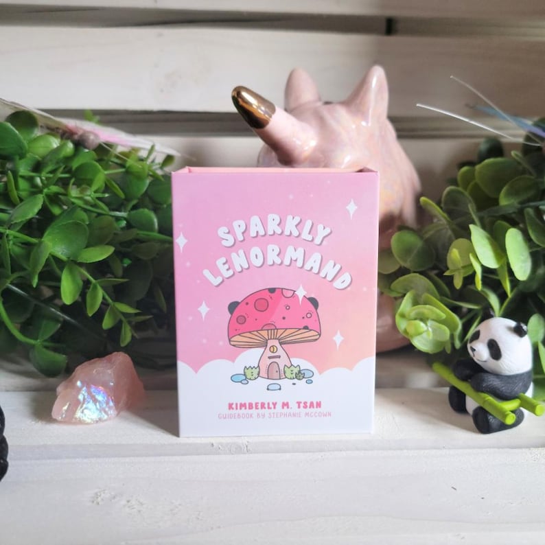 May include: A pink box with the text "Sparkly Lenormand" and a cartoon mushroom house with a pink roof. The text "Kimberly M. Tsan" and "Guidebook by Stephanie McCown" are also on the box.