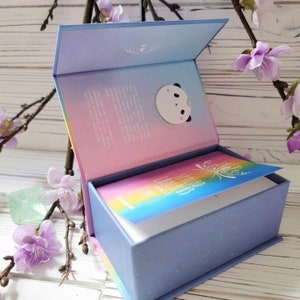 K&ouml;nnte beinhalten: Eine blaue Box mit einem Regenbogen-Design und dem Text "Spread Creativity" auf der Vorderseite. Die Box ist ge&ouml;ffnet und enth&auml;lt ein Kartenspiel mit einem Regenbogen-Design und einer Panda-Illustration auf der Innenseite des Deckels.