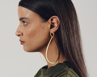 May include: A pair of large, gold-toned teardrop-shaped earrings with a smooth, polished finish. The earrings feature a slender, open-loop design that hangs vertically from the earlobe, creating a long, minimalist silhouette against the side profile.
