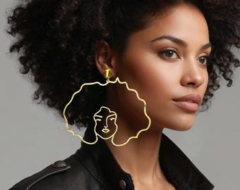 May include: A large gold-toned earring featuring a continuous wire outline of a face with curly hair, worn on an ear. The earring has a thin, metallic frame that mimics the shape of a voluminous afro hairstyle and a simple facial profile.