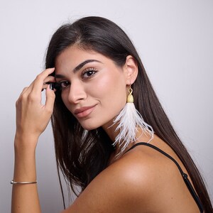 May include: A woman with long dark hair wearing a black tank top and white feather earrings. She is wearing a silver bracelet on her wrist and has her hand on her head.