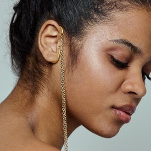 May include: A woman with dark hair pulled back in a bun wears a gold chain ear cuff. The chain hangs down the side of her neck.