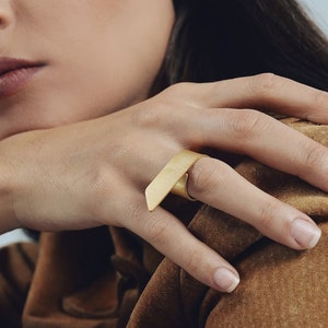 May include: A gold-toned ring featuring a wide, flat, rectangular bar design that sits horizontally across the finger. The metal has a smooth, matte finish, and the band is circular, providing a clean and simple aesthetic for the hand.