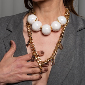 May include: A gold-toned chain necklace featuring five large, smooth white spherical beads and several small, rectangular dark-colored charms with gold-toned borders, worn over a grey blazer with notched lapels and a structured shoulder design.