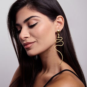 Peut inclure: Une femme aux longs cheveux noirs portant un haut noir et des boucles d'oreilles pendantes dorées avec un design abstrait unique.