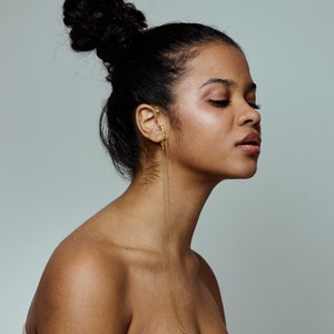 May include: A woman with dark hair styled in a bun, wearing a gold ear cuff with a long chain. She has her eyes closed and is facing to the right. The background is a solid, light gray color.