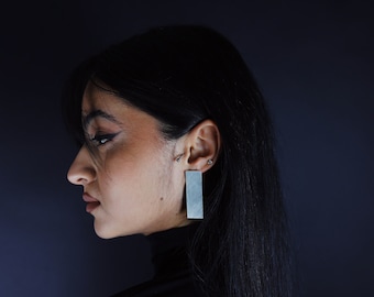May include: A silver-toned rectangular drop earring with a brushed metal finish. The earring features a long, flat, vertical bar shape that hangs from the earlobe, displaying a matte, textured surface that reflects light across its length.