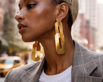 May include: A pair of gold-toned drop earrings featuring two elongated, rectangular chain links connected vertically. The earrings have a smooth, matte metallic finish and are worn with a grey plaid blazer, a white t-shirt, and blue denim jeans.