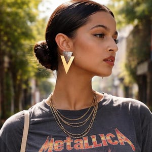 Puede incluir: Una mujer con un top corto gris con la palabra "Metallica" en letras naranjas y un gráfico de calavera. También lleva collares dorados en capas y pendientes triangulares grandes dorados y plateados.