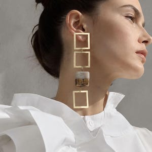May include: A long drop earring featuring two gold-toned open square frames and a central square block with a marbled white and brown pattern. The earring hangs vertically, with the gold squares positioned at the top and bottom of the block.