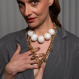 May include: A multi-layered necklace featuring a thick gold-tone chain and a strand of large, iridescent white spherical beads. The gold chain is adorned with small, rectangular charms featuring a dark purple center with a gold-tone eye motif.
