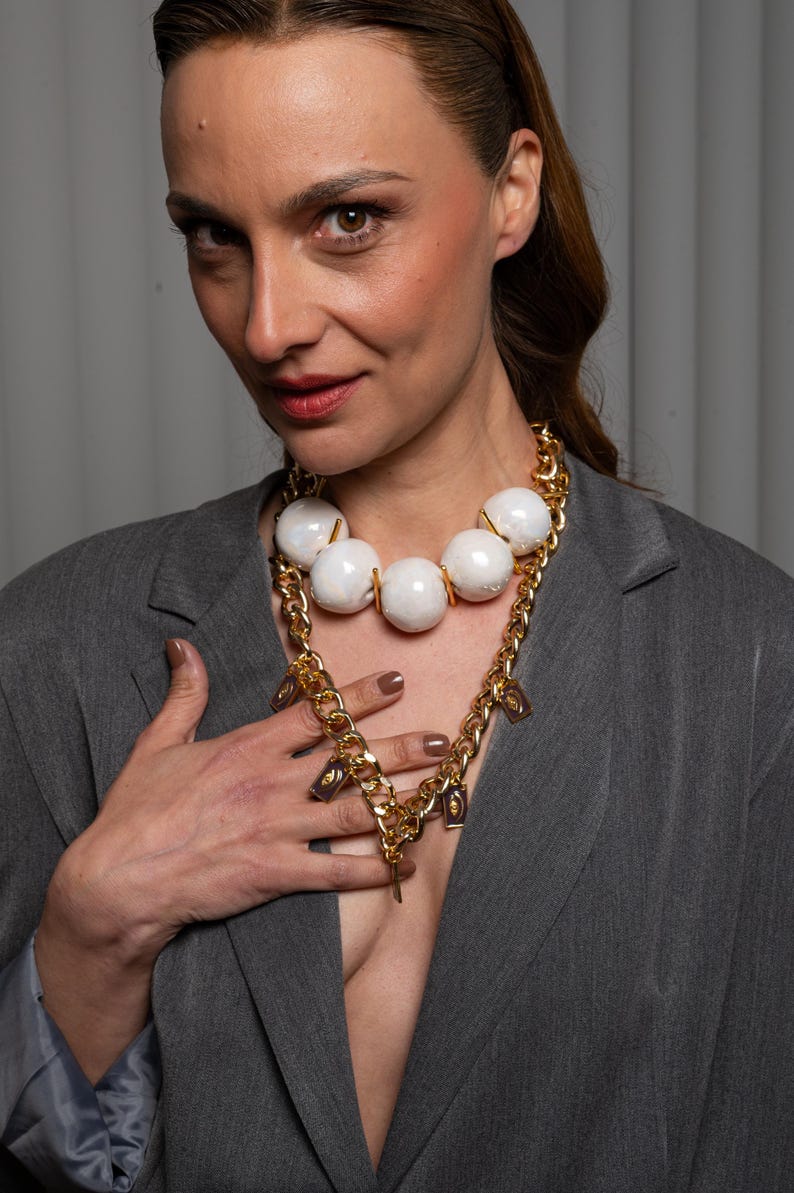 May include: A multi-layered necklace featuring a thick gold-tone chain and a strand of large, iridescent white spherical beads. The gold chain is adorned with small, rectangular charms featuring a dark purple center with a gold-tone eye motif.