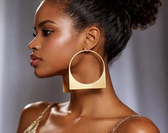 May include: A large gold-toned earring featuring a geometric design that combines a circular upper section with a flat, horizontal base. The metal has a smooth, matte finish and is worn as a statement piece on the earlobe, showcasing a bold shape.