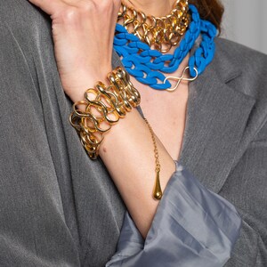 May include: A gold chain bracelet with a dangling teardrop charm. The bracelet is made of thick, chunky links and is worn on a person's wrist. The person is also wearing a layered necklace with a blue plastic chain and a gold chain.