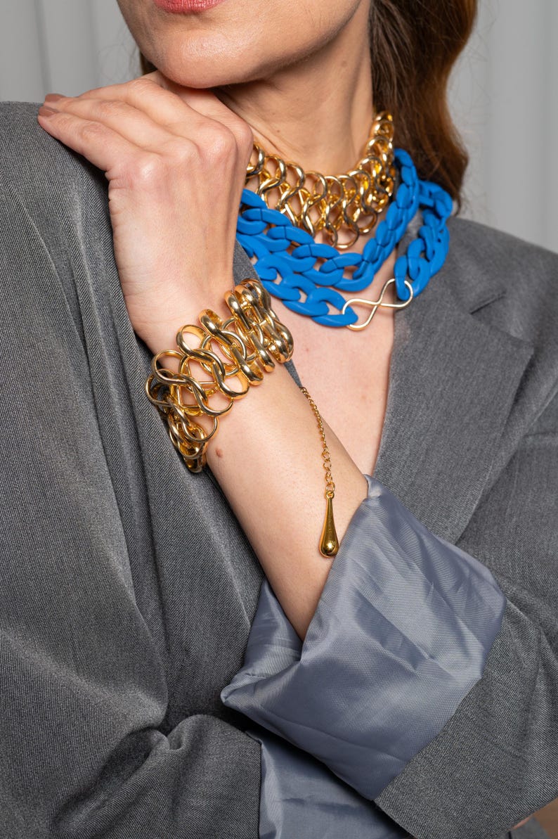 May include: A gold chain bracelet with a dangling teardrop charm. The bracelet is made of thick, chunky links and is worn on a person's wrist. The person is also wearing a layered necklace with a blue plastic chain and a gold chain.