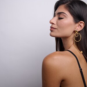 May include: A woman with long dark hair wearing a black vest top and gold hoop earrings. The earrings are large and have a unique design.