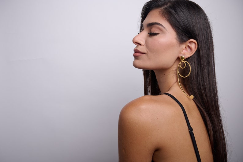 May include: A woman with long dark hair wearing a black vest top and gold hoop earrings. The earrings are large and have a unique design.