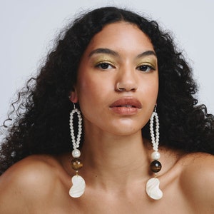 May include: A woman with long, curly black hair wearing a pair of long, pearl and tiger's eye drop earrings with a large white shell charm at the bottom.