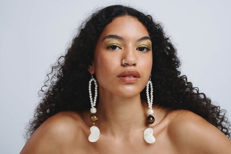 May include: A woman with long, curly black hair wearing a pair of long, pearl and tiger's eye drop earrings with a large white shell charm at the bottom.