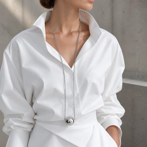 Puede incluir: Un collar largo plateado con un colgante esférico grande. El collar se lleva con una camisa blanca con cuello y una falda envolvente blanca. El colgante es una bola plateada brillante y reflectante.