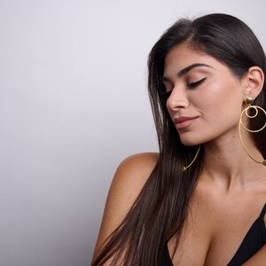May include: A woman with long dark hair wearing a black vest top and large gold hoop earrings with a dangling circle.