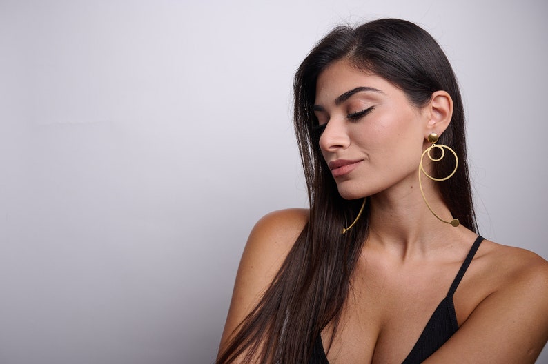 May include: A woman with long dark hair wearing a black vest top and large gold hoop earrings with a dangling circle.
