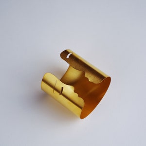 May include: A gold-toned metal cuff bracelet featuring a wide, open-ended band design. The edges of the cuff are cut into a distinct, irregular silhouette that resembles the profile of a human face, with small curved cutouts forming eye details.